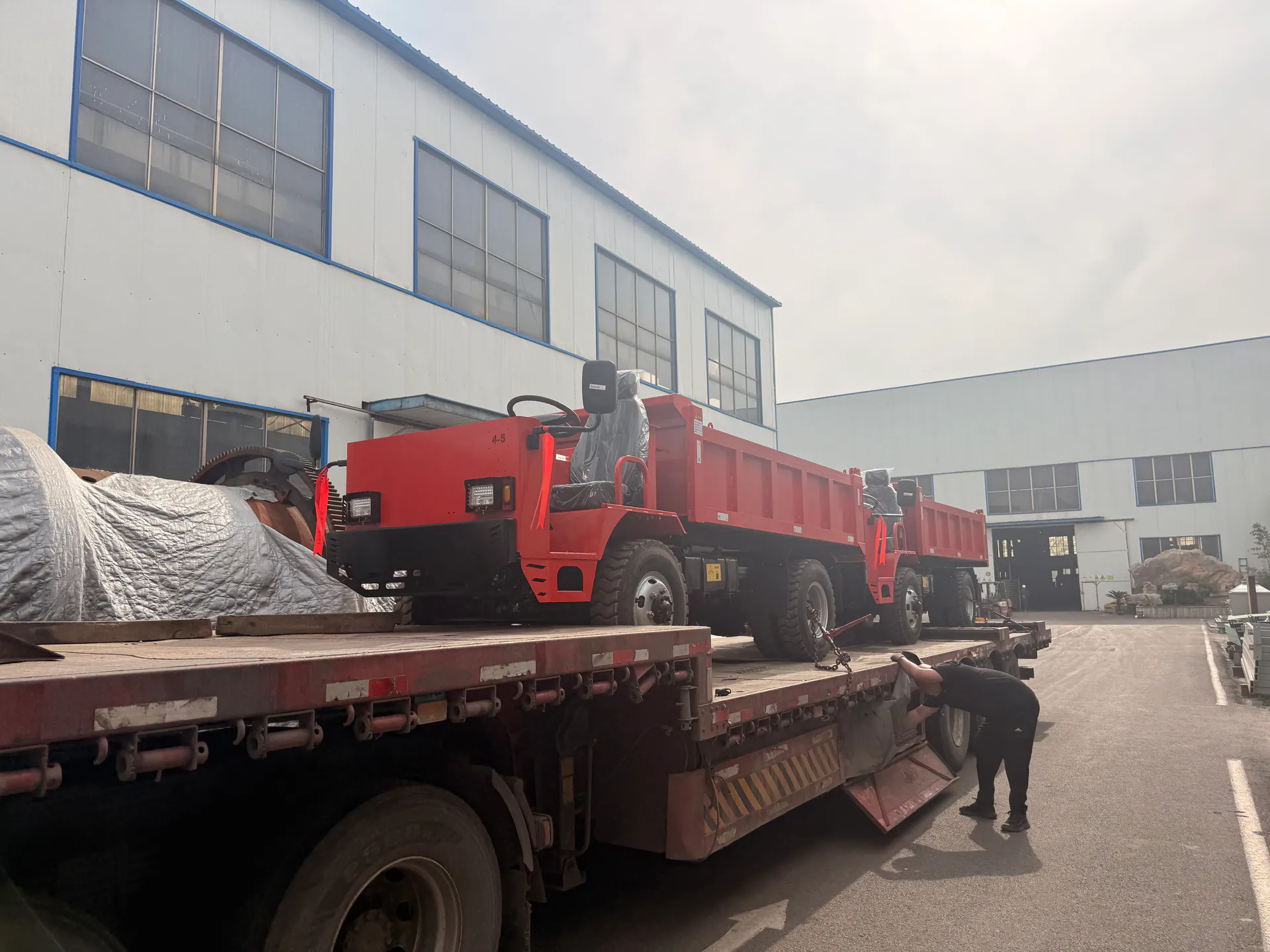 A person beside a red mining truck positioned on a transport platform outside a workshop.